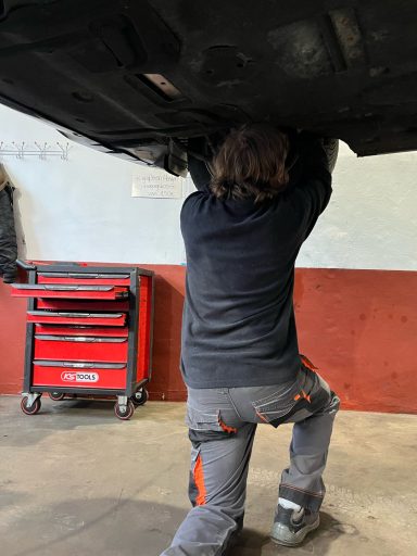 Person arbeitet unter einem Auto, greift nach einer Stelle am Fahrzeug. Werkzeugkiste im Hintergrund.