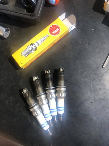 Vier Zündkerzen mit blauen und silbernen Akzenten auf einem Tisch, daneben eine Verpackung.